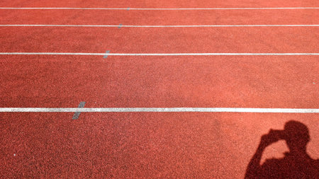 Photo of a red athletic track with white lane markings in the sunlight. A clear human shadow appears...