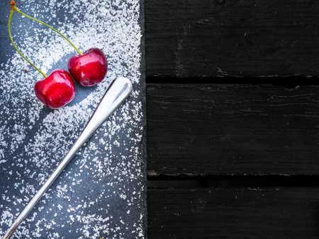 Two fresh red cherries on a dessert board sprinkled with sugar and served with a silver spoon viewed from above with copy spaceの写真素材
