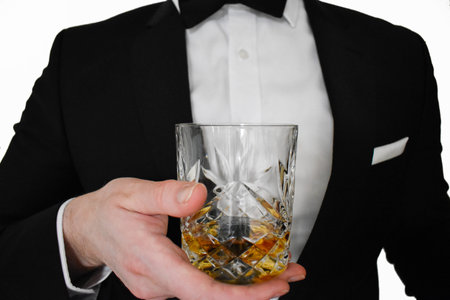 A close up view of a man in a black tuxedo offering a glass of whiskey