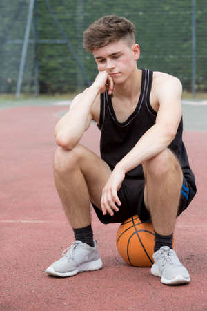 Teenage boy sitting on a basketball on a court looking sadの写真素材