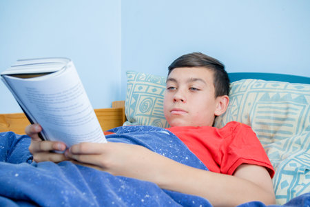 Caucasian teenage boy reading a book laying on a bed