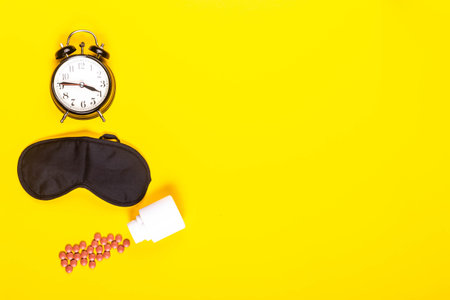 Sleep concept showing an eye mask, alarm clock and sleeping pills on a yellow background