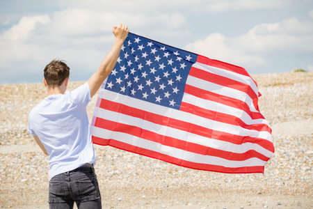 Young adult caucasian male holding on a beach holding the flag of The United States