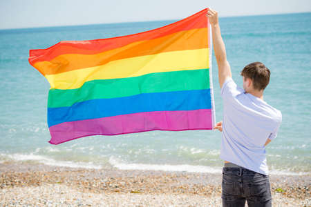 Young adult caucasian male holding on a beach holding the Pride flagの写真素材