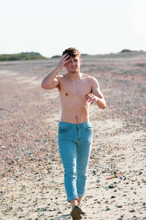 Young caucasian shirtless man wearing blue jeans walking on a beach on a warm summerâs afternoonの写真素材