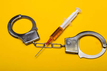 Illegal drug crisis concept showing a full syringe and handcuffs on a yellow backgroundの写真素材