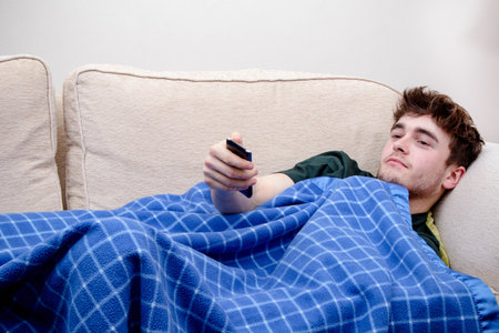 A young adult man laying on a sofa, using a remote, wrapped in a blanket