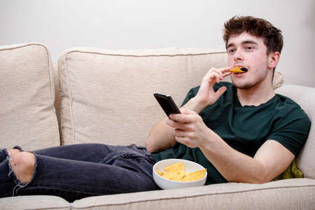 A young adult man laying on a sofa watching TV and eating snacksの写真素材
