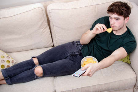 A young adult man laying on a sofa watching TV and eating snacksの写真素材