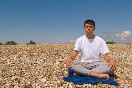 A Caucasian teenage boy meditating on a stony beach with his hands resting on his kneesの写真素材