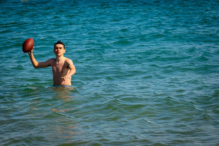 A 18 year old Caucasian shirtless teenage boy in swimwear playing with an American Football in the seaの写真素材