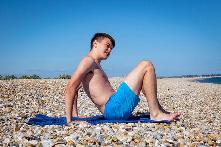 A 18 year old Caucasian shirtless teenage boy sitting on a stony beach