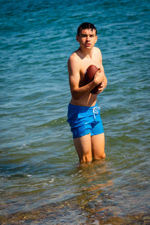 A 18 year old Caucasian shirtless teenage boy in swimwear playing with an American Football in the seaの写真素材