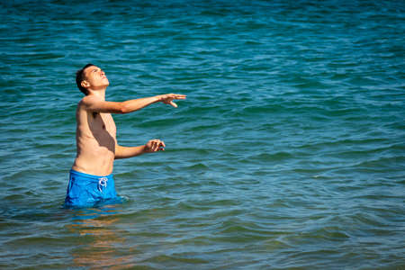 A 18 year old Caucasian shirtless teenage boy in swimwear playing with an American Football in the seaの写真素材