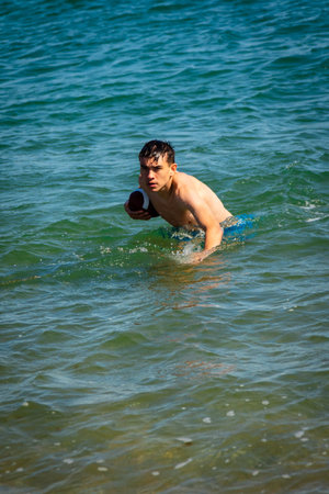 A 18 year old Caucasian shirtless teenage boy in swimwear playing with an American Football in the seaの写真素材