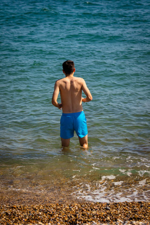 A 18 year old Caucasian shirtless teenage boy in swimwear walking into the sea