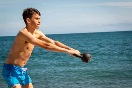 A 18 year old Caucasian shirtless teenage boy on a beach exercising with a kettlebell weightの写真素材