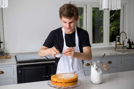 A teenage boy in a domestic kitchen putting icing sugar on a cakeの写真素材
