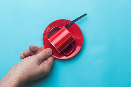 An Espresso coffee cup, spoon and saucer held by a hand on a blue background