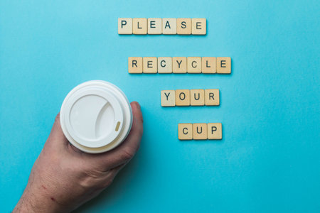 A paper coffee cup amd lid with a sign reading Please Recycle Your Cup held by a human hand on a blue backgroundの写真素材