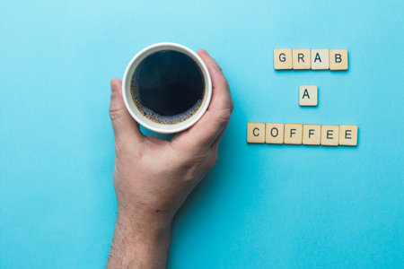 An open paper cup of black cofffee held by a hand on a blue background with a sign reading grab a coffeeの写真素材