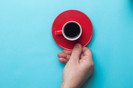 An Espresso cup of black coffee on a saucer being held by a hand on a blue back groundの写真素材