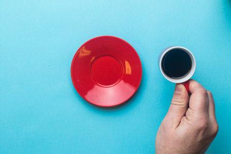 An Espresso cup of black coffee being held by a hand on a blue background with a saucerの写真素材