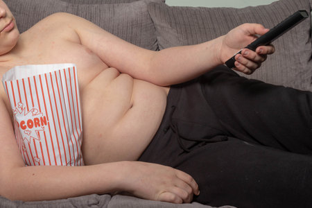 An overweight caucasian teenage boy laying on a sofa watching tv with popcornの写真素材
