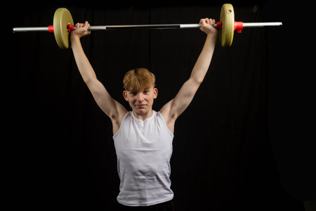 Portrait of a 14 year old caucasian teenage boy wearing a sleeveless vest lifting a barbell against a black background boyの写真素材