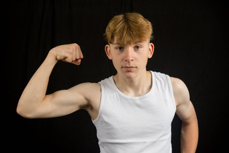 Portrait of a 14 year old caucasian teenage boy wearing a sleeveless vest flexing his bicep against...