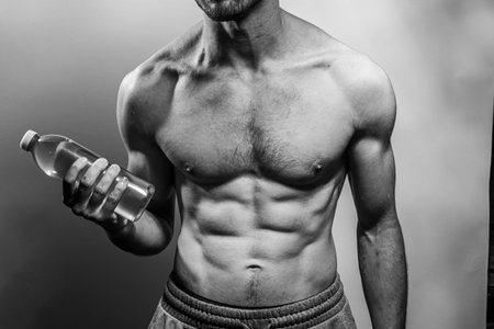 A young male body holding a water bottle, with well-defined abs and chest. Model with shirtless torso. Concept of body care, male health, fitness, youth, masculinity, health, weightloss, bodybuildingの写真素材