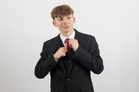 Smiling Sixteen year old teenage boy wearing a suit adjsuting his tie, studio shot against a white backgroundの写真素材