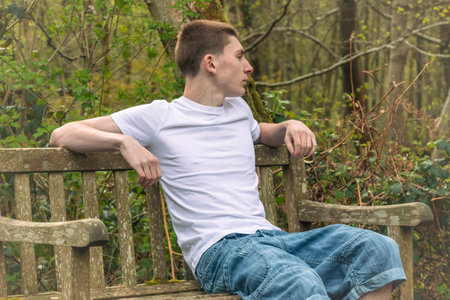 Teenage boy sititng on a bench in a public park, wearing a white t-shirt and blue denimの写真素材
