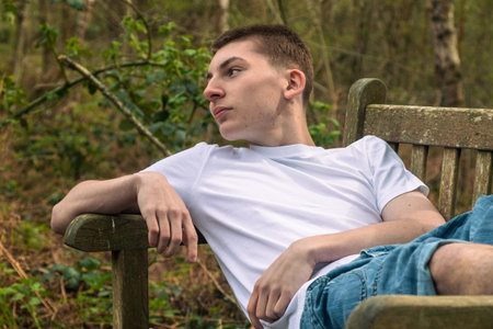 Happy Teenage boy laying on a bench in a public park, wearing a white t-shirt and blue denimの写真素材