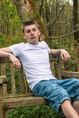 Teenage boy sitting on a bench in a public park, wearing a white t-shirt and blue denimの写真素材