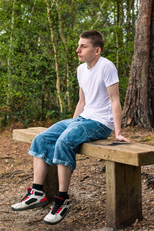 Teenage boy sititng on a bench in a public park, wearing a white t-shirt and blue denimの写真素材