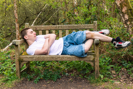 Teenage boy sleeping on a bench in a public park, wearing a white t-shirt and blue denimの写真素材