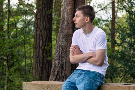 Teenage boy sitting on a bench in a park, wearing a white t-shirt and blue denimの写真素材