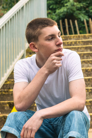 Serious Teenage boy with his chin on his hand, in white t-shirt and blue denim sitting on stairs outside in a public parkの写真素材