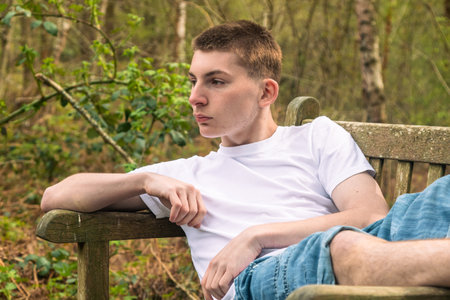 Teenage boy sititng on a bench in a public park, wearing a white t-shirt and blue denimの写真素材