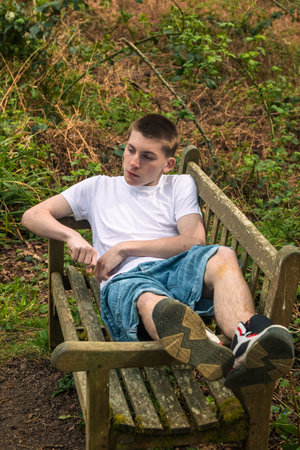 Teenage boy laying on a bench in a public park, wearing a white t-shirt and blue denimの写真素材