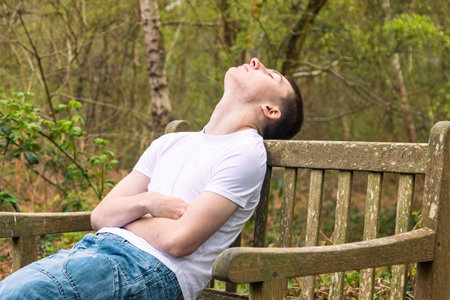 Teenage boy sleeping on a bench in a public park, wearing a white t-shirt and blue denimの写真素材