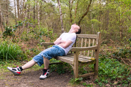 Teenage boy sleeping on a bench in a public park, wearing a white t-shirt and blue denimの写真素材