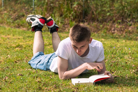 Teenage boy reading a book in the sun in a public park. Wearing a white T-shirt and blue denimの写真素材