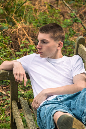 Teenage boy laying on a bench in a public park, wearing a white t-shirt and blue denimの写真素材