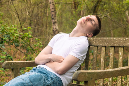 Teenage boy sleeping on a bench in a public park, wearing a white t-shirt and blue denimの写真素材