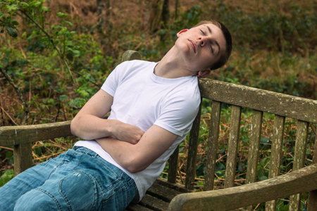 Teenage boy sleeping on a bench in a public park, wearing a white t-shirt and blue denimの写真素材