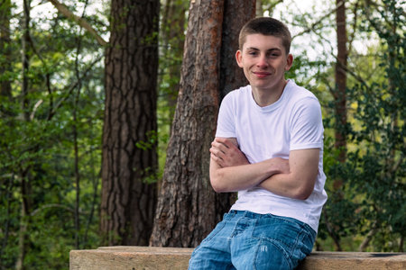 Teenage boy sititng on a bench in a public park, wearing a white t-shirt and blue denimの写真素材