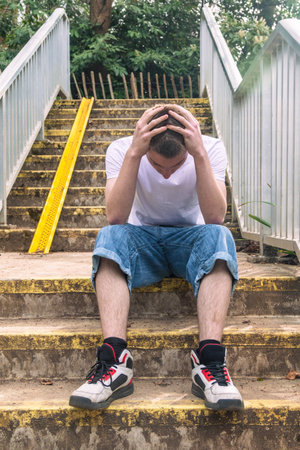 Depressed Teenage boy with his head in his hands, in white t-shirt and blue denim sitting on stairs outside in a public parkの写真素材