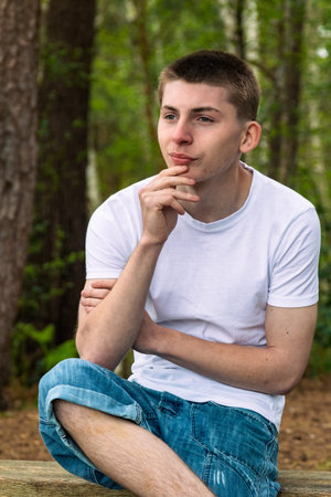 Happy Teenage boy sititng on a bench in a public park, wearing a white t-shirt and blue denimの写真素材